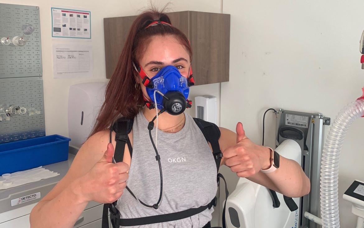 Ava participating in a validation test with a piece of equipment that measures ardiorespiratory fitness levels during the leg and arm cycling tests. She is giving two thumbs up.